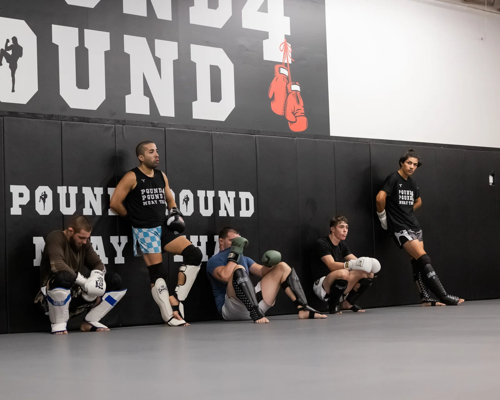 Pound 4 Pound Muay Thai students resting between rounds against the wall inside the Littleton training gym